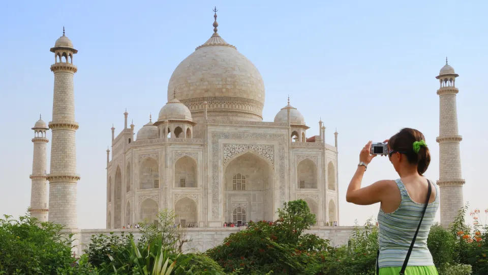Visitor photographing the Taj Mahal from the gardens, with the central dome and minarets aligned against a clear sky