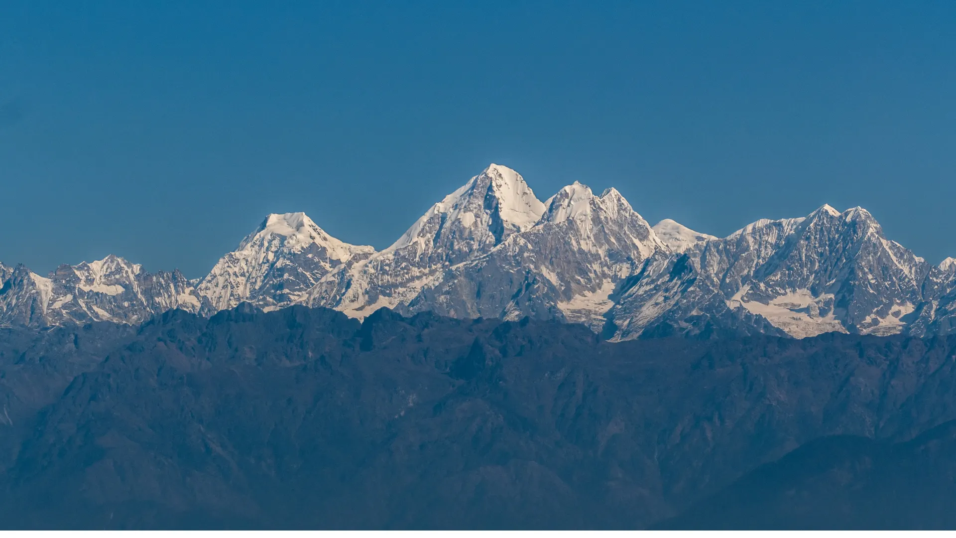 The snow-covered Himalayan range in clear dawn light seen across forested foothills from the Nagarkot ridge