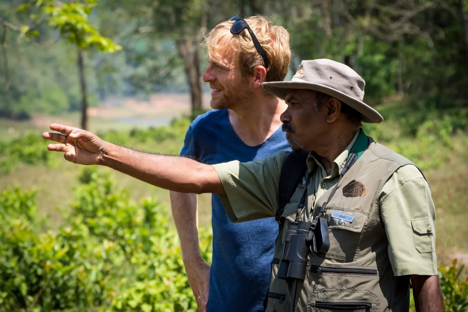 A nature guide pointing toward the forest while another person stands beside them in a lush green landscape
