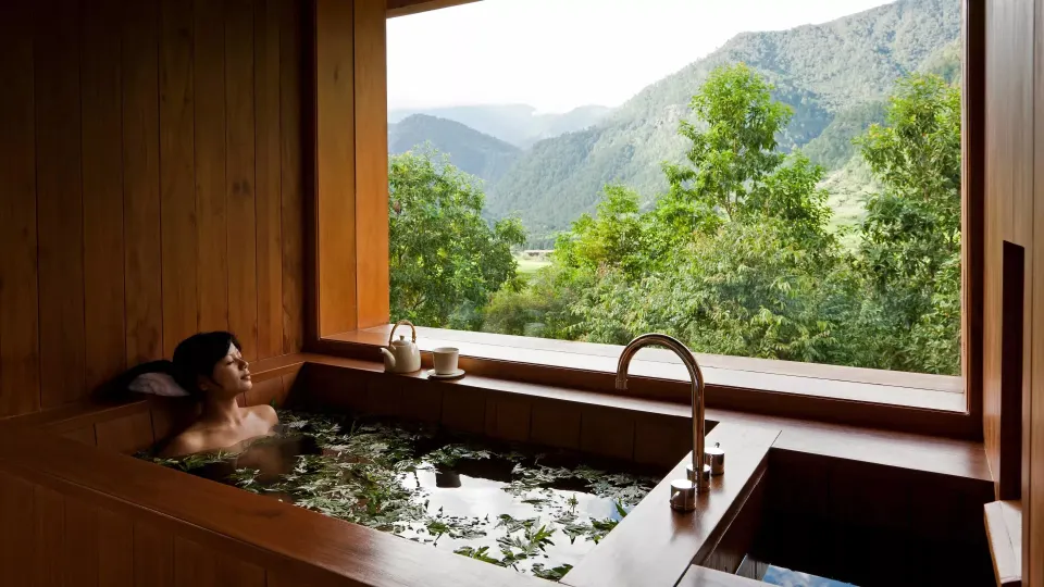 Person soaking in a wooden herbal bath with green mountain views through a window