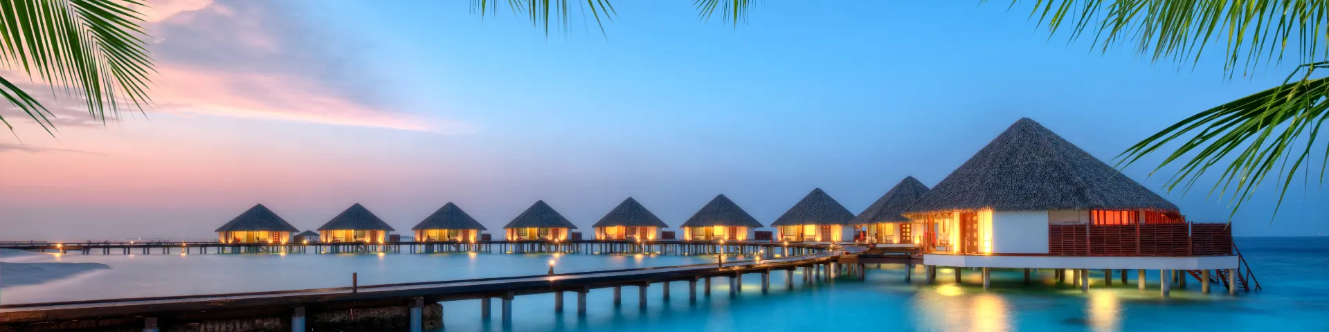 Overwater villas at dusk with a calm lagoon, soft evening light, and palm fronds framing the scene